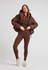 Brązowa puchowa kurtka z kapturem, z pikowanym wzorem; brązowy dopasowany top i legginsy, uzupełnione srebrnymi butami sportowymi.