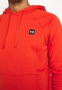 Felpa rossa realizzata in tessuto morbido, con tasca a canguro, cappuccio con lacci e un piccolo logo nero sul petto.