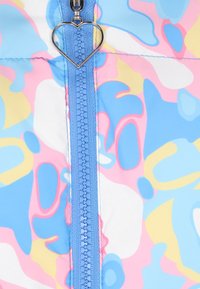 Farverig lynlåsjakke med en blå lynlås og hjerteformet trækflig. Stoffet viser et multifarvet abstrakt mønster med pink, blå og gul.