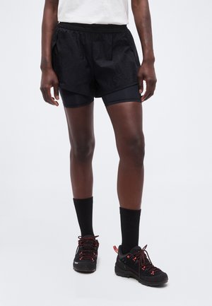 Picioare purtând pantaloni scurți negri pentru alergare stratificați, șosete negre și pantofi negri de drumeție cu șireturi roșii, stând pe un fundal alb simplu.