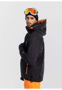 Giacca da sci nera con una fodera e dettagli arancioni, dotata di cappuccio, tasche con zip e una vestibilità sportiva, abbinata a occhiali da sci.