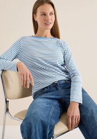 Haut à manches longues rayé en bleu clair et beige, porté avec un jean bleu. Le mannequin est assis sur une chaise, mettant en valeur la coupe décontractée de la chemise.