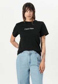 Zwarte katoenen T-shirt met korte mouwen, met in het midden het witte "Calvin Klein" logo. Ggedragen met lichtblauwe high-waisted jeans.