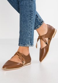 Ruske nahodne cipele od kože s šiljastim vrhom, koje imaju čvorni detalj i metalni naglasak; nose se s plavim skraćenim trapericama na bijeloj površini.