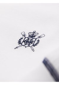 Tissu blanc avec un blason brodé bleu marine comprenant une couronne, un bouclier, des avirons croisés et des branches de laurier.