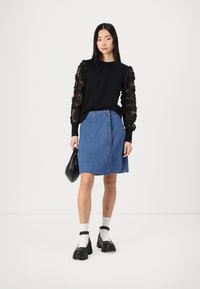 Schwarzer Pullover mit transparenten floralen Ärmeln, kombiniert mit einem hellblauen Denimrock. Das Model trägt schwarze klobige Schuhe und eine kleine schwarze Tasche.