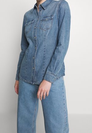 Femme portant une chemise en denim bleu clair boutonnée avec poches poitrine et jean large assorti, sur un fond uni.