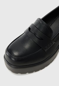 Sapatos de couro pretos tipo loafer com um acabamento suave, apresentando uma faixa larga no peito do pé e uma sola grossa e texturizada.