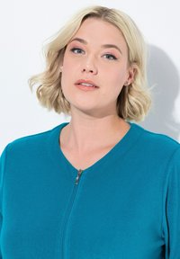 Giacchetto teal con zip a tutta lunghezza e scollatura rotonda, realizzato in tessuto morbido. Presenta una zip liscia e dettagli di cucitura sulle spalle.