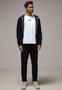 Felpa nera con zip in tessuto testurizzato e dettagli verde, t-shirt bianca, pantaloni neri e sneaker bianche con suole beige.