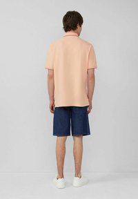 Pfirsichfarbenes Poloshirt mit kurzen Ärmeln und kontrastierenden Kragenakzenten, kombiniert mit knielangen blauen Jeansshorts und weißen Sneakers.
