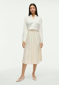 Nainen seisoo päällä valkoinen pitkähiha blouse, pleated vaaleankerman värinen midi-hame ja nude matalakorkoiset suutret kengät tasaisella taustalla.