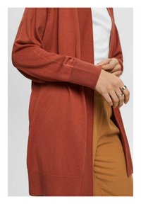 Cardigan couleur rouille avec bords côtelés, en maille légère, design à ouverture frontale et deux poches latérales. Porté sur un haut blanc.