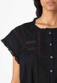 Blusa nera con dettagli in pizzo, maniche corte, scollatura rotonda e bottoni frontali. Presenta motivi testurizzati e delicati bordi a scallop.