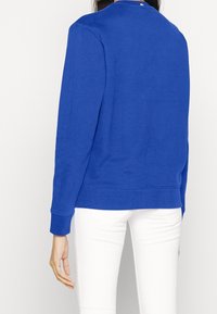 Modrý svetr s dlouhými rukávy a kulatým výstřihem, vyrobený z texturované látky. Má rovný lem a žebrované manžety. Bílé kalhoty viditelné dole.