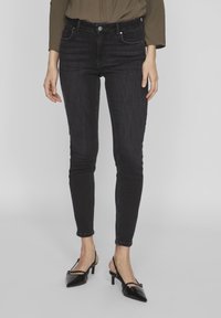 Jeans negros ajustados con cintura alta, bolsillos frontales y detalles de costura sutiles. Combinados con tacones negros de punta afilada.