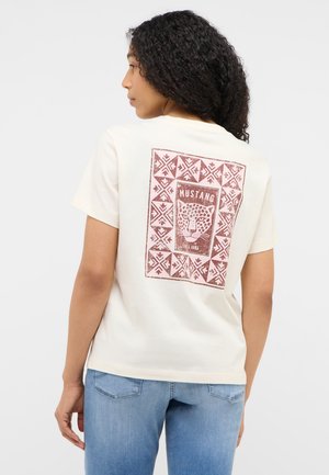 Woman with curly hair wearing white T-shirt with brown leopard print and "Mustang Since 1932" text, paired with blue jeans, facing sideways.