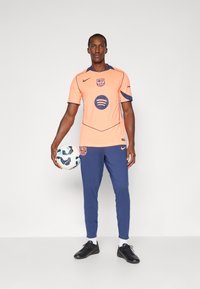 Het voetbalshirt voor heren heeft een fel oranje trui met blauwe accenten, een logo en het Spotify-embleem; gecombineerd met marineblauwe broek en zwarte voetbalschoenen.