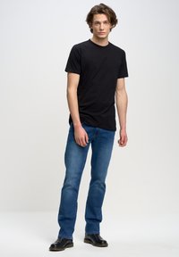 Camiseta de algodón negra, de cuello redondo, combinada con jeans de mezclilla azules y botas de cuero negras. El modelo se encuentra en un fondo neutro.