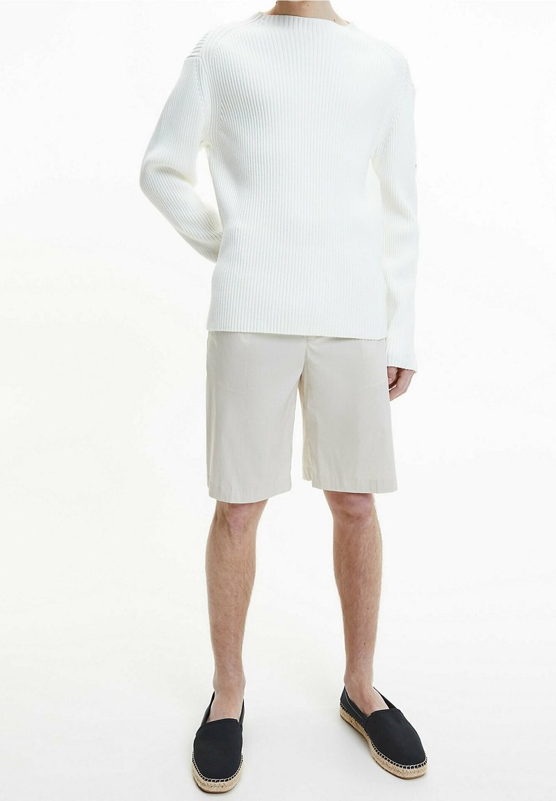 Une personne se tient debout, vêtue d'un pull à manches longues blanc côtelé, d'un short beige arrivant aux genoux et d'espadrilles noires, sur un fond blanc.