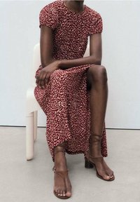 Personne assise portant une robe à motifs rouges et blancs et des sandales à talons bruns à lanières sur un fond blanc uni.