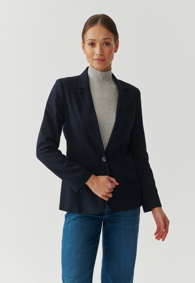 Blazer navy en tissu léger avec une fermeture à un bouton, revers crantés et deux poches. Associé à un col roulé gris et à un jean.