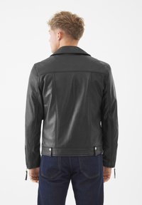 Mann mit kurzen lockigen Haaren, der eine schwarze Lederjacke und dunkelblaue Jeans trägt, von hinten vor einem weißen Hintergrund gezeigt.