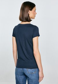 Tumši zils, īsām piedurknēm T-krekls ar apaļu kakla izgriezumu. Izgatavots no mīksta auduma, tam ir pieguļošs griezums un gluda tekstūra, kombinēts ar ziliem džinsiem.