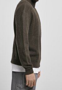 Maglione lavorato a maglia marrone con colletto alto e polsini a costine, caratterizzato da una finitura testurizzata. Indossato sopra una camicia bianca e pantaloni grigi.