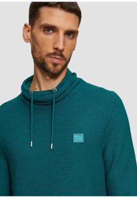 Pullover turchese con collo alto, dotato di cordoncino e piccolo logo applicato. Il tessuto ha un aspetto testurizzato e a coste.