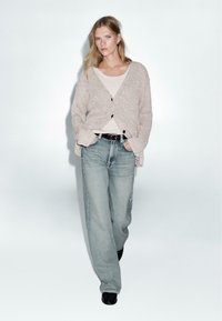 Světlý béžový pletený kardigan s V výstřihem a propínacím předním dílem, nošený přes smetanový top a spárovaný se světle modrými kalhotami s širokými nohavicemi. Černé boty.