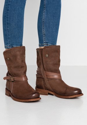 Bottes en cuir marron mi-mollet avec fermeture éclair latérale et sangle, portées avec un jean skinny bleu sur un sol blanc et un fond clair.
