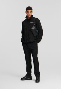 Schwarze Außenjacke mit hohem Kragen, kombiniert mit passenden Hosen. Eine strukturierte schwarze Umhängetasche hängt von der Schulter. Schwarze Schuhe.
