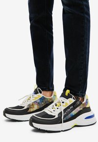 Zapatillas deportivas multicolores con parte superior de malla, suela blanca y negra, detalles en amarillo y patrón reflectante. Cordones y cierre de cremallera.