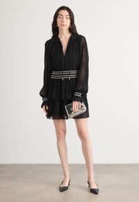 Black pleated dress with a deep V-neck, long sleeves, and a silver-accented waistband. Silver clutch and black pointed heels complete the look.