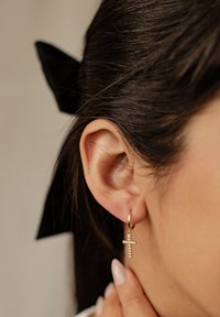 Goldene Creolen mit einem kleinen Kreuzanhänger, der klare Steine zeigt, am Ohrläppchen getragen, passend zu dunklem Haar und einem schwarzen Schleifenaccessoire.