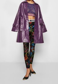 Un lungo cappotto viola lucido sopra un top in rete blu a maniche corte, abbinato a leggings neri con motivi barocchi colorati e tacchi neri.