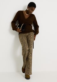 Suéter marrón envolvente, pantalones acampanados con estampado de leopardo y botas oscuras. Los accesorios incluyen un bolso negro con detalle de cadena dorada y un collar.