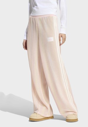 Osoba nosząca jasno różowe, szerokie teksturowane spodnie z bocznymi paskami i naszywką z logo, w połączeniu z beżowymi płaskimi butami i białą, długorękawową bluzką.