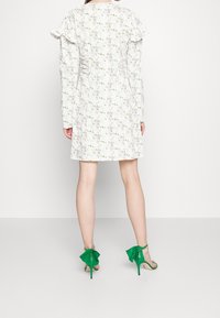 Vestido floral blanco con mangas largas y hombros con volantes, combinado con sandalias de tacón verdes que presentan acentos en forma de hojas.