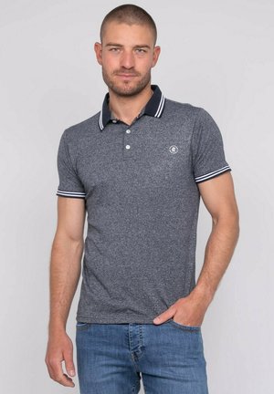 Homme aux cheveux courts et barbe, portant un polo gris avec col et manches rayés, jean bleu, debout avec une main dans la poche.