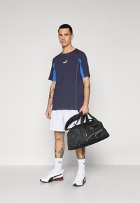 Donkerblauw T-shirt met korte mouwen en blauwe accenten, witte shorts, zwarte gymtas en witte sneakers met rode details. Tattoos zichtbaar op de benen.