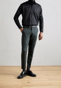 Zwart overhemd met knopen, gecombineerd met slim fit donkergroene broek en gepolijste zwarte geklede schoenen. De houten vloer op de achtergrond benadrukt de outfit.
