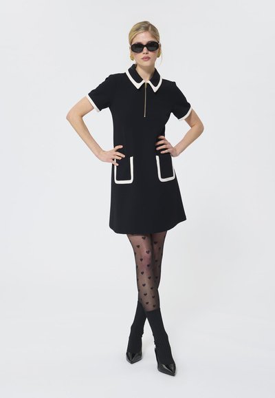 Vestido negro con cuello blanco y bolsillos, de manga corta y con cremallera. Combinado con medias negras con estampado de corazones y zapatos de tacón alto.