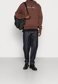 Brun huvtröja med vit logotyp, kombinerad med mörkblå denimbyxor i lös passform och svarta skor. Inkluderar en svart väska över axeln.