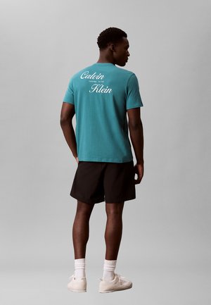 Mann trägt ein türkisfarbenes Calvin Klein T-Shirt, schwarze Shorts, weiße Socken und weiße Sneaker und steht mit dem Rücken zur Kamera.