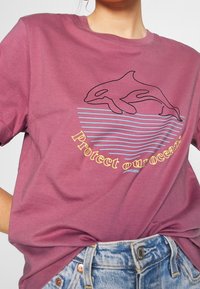 Kastanjebrun bomull t-shirt med en svart kontur av en delfin och texten "Skydda våra hav" i gult, med blå horisontella ränder.