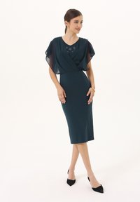 Robe teal avec un drapé fluide en voile, silhouette ajustée, décolleté en V, ornements sur la poitrine, longueur genou, associée à des talons noirs.
