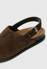 Καφέ σουέντ σανδάλι με ανοιχτή πλάτη, ρυθμιζόμενο λουράκι με ασημένια αγκράφα και επίπεδη μαύρη σόλα με μπεζ εσωτερική επένδυση.
