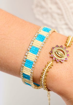Mehrreihiges Armband mit blauen und cremefarbenen Perlen, goldfarbenen Akzenten und einem dekorativen Sonnenanhänger mit pinken und klaren Steinen.